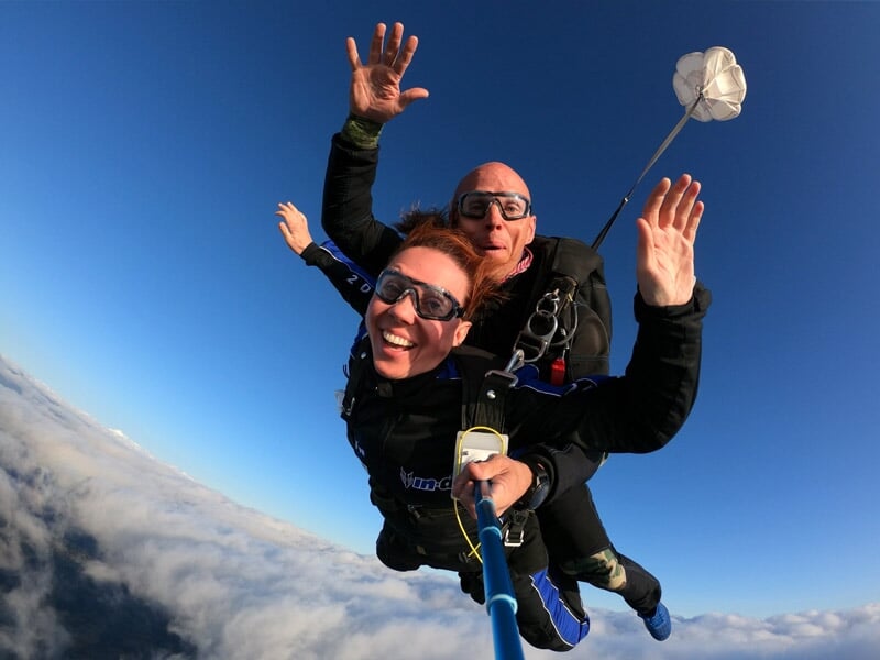 Saut en tandem avec une femme souriante et un instructeur, parachute déployé en arrière-plan, entourés d’un ciel bleu et de nuages