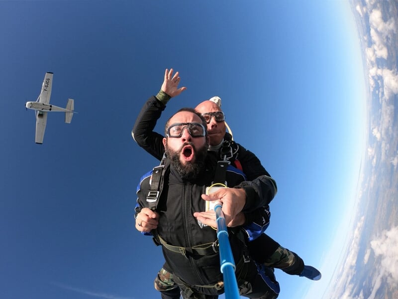 Saut en parachute en tandem avec un instructeur, le visage d’un homme exprimant l’excitation en plein vol, un avion visible en arrière-plan dans un ciel dégagé