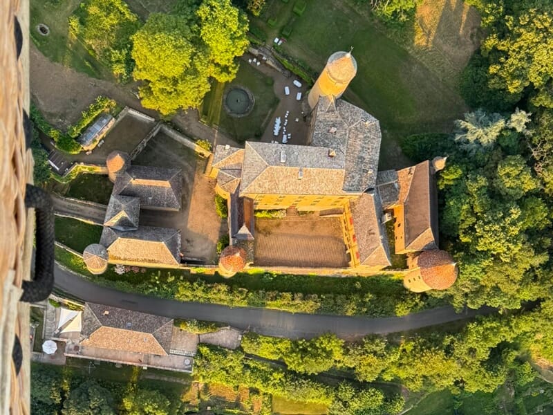Vue aérienne d'un château entouré d'arbres verts et d'un chemin goudronné, prise depuis une montgolfière.