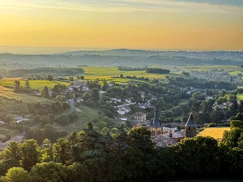 Paysage vallonné avec village, champs et arbres sous un ciel au coucher du soleil doré.