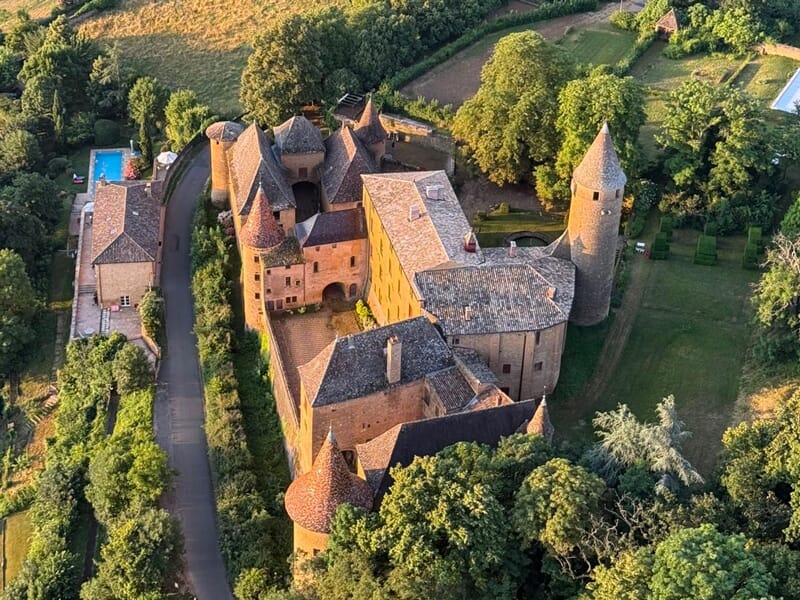 Château médiéval avec tours rondes entouré de verdure et d'un chemin sinueux, vu d'en haut.