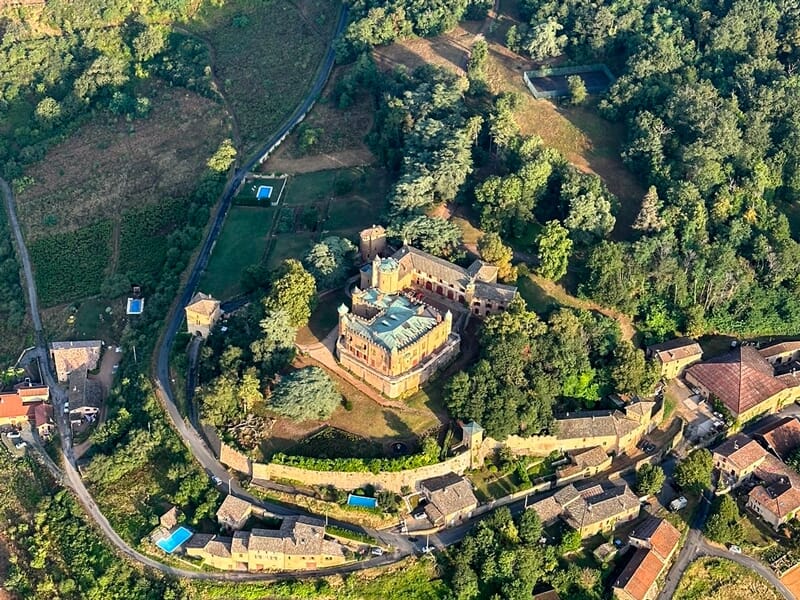 Vue aérienne d'un château fortifié entouré d'arbres et de maisons, avec une route en courbe.