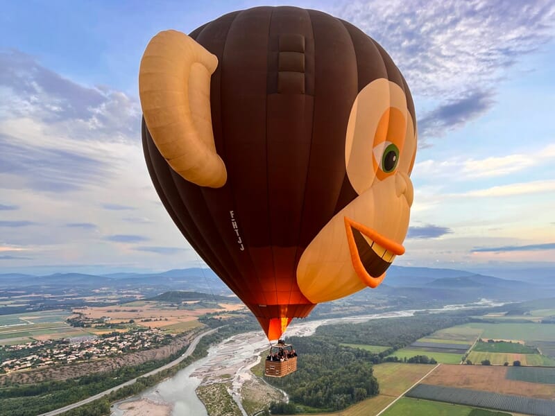 Montgolfière en forme de tête de singe survolant une rivière et des champs au coucher du soleil.