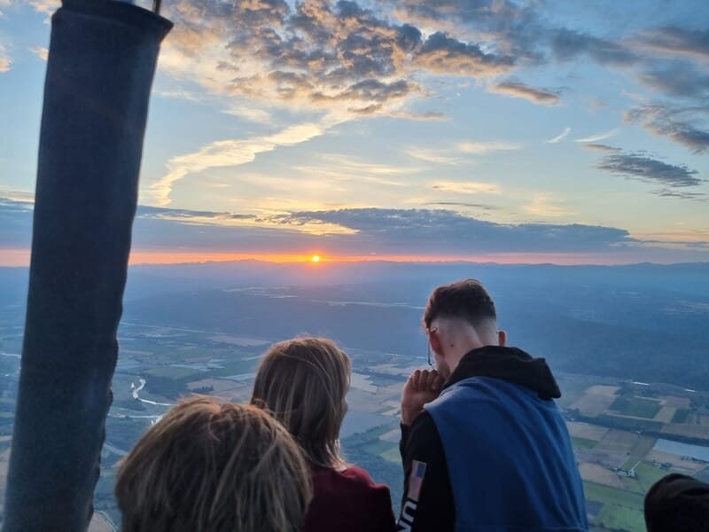 Trois personnes dans une montgolfière regardant le coucher de soleil sur un paysage vallonné et une rivière.