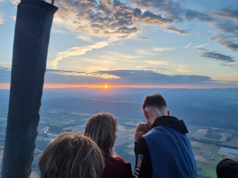 Trois personnes dans une montgolfière regardant le coucher de soleil sur un paysage vallonné et une rivière.