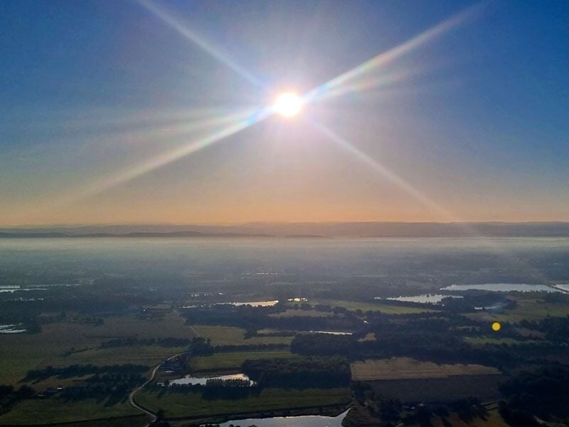 Vue aérienne au lever du soleil sur un paysage rural avec champs, forêts et plans d'eau sous un ciel clair.