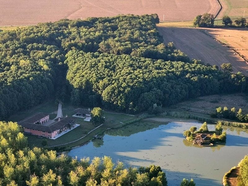 Vue aérienne d'un domaine avec un bâtiment, un étang et une petite île entourés de forêts et champs agricoles.