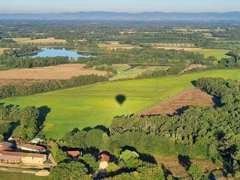 Vue aérienne d'un champ vert avec une ombre de montgolfière, des arbres et des bâtiments en bordure.
