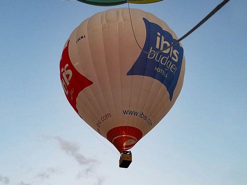 Montgolfière blanche avec logos ibis budget en vol, suspendue dans un ciel clair au-dessus du sol.