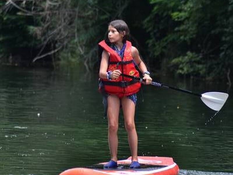 Jeune fille debout sur un paddle, pagayant sur une rivière calme entourée de végétation dense.