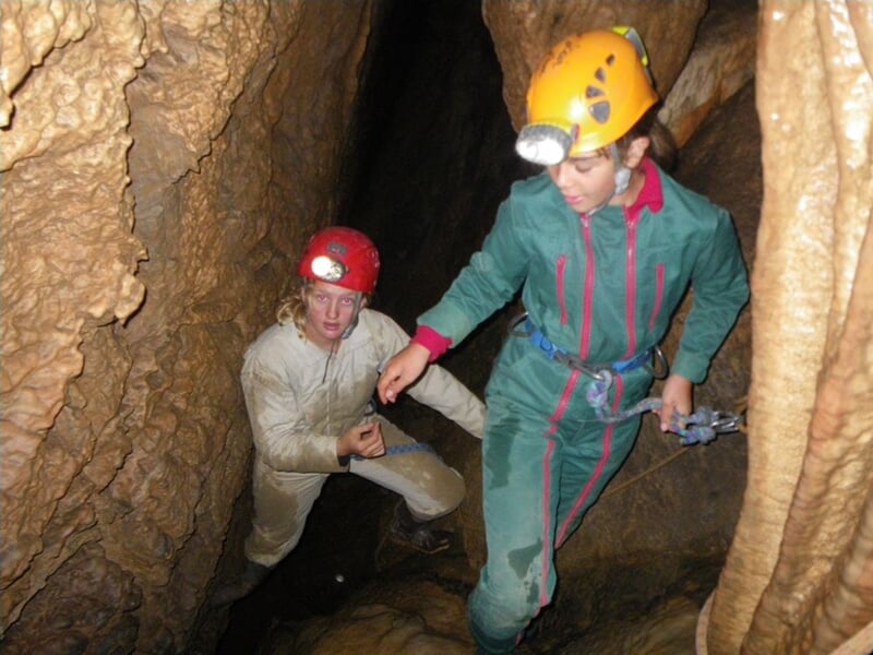 Deux spéléologues en combinaison et casque explorent une grotte étroite avec des parois rocheuses.