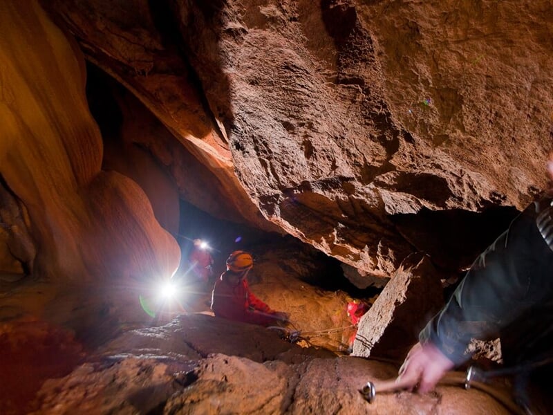 Explorateurs en spéléologie progressant dans une grotte sombre avec éclairage artificiel et roches.