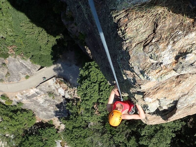 Escaladeur en casque blanc grimpe une paroi rocheuse abrupte dans un cadre naturel verdoyant.