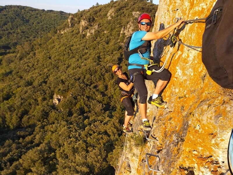 Couple équipé de casques et harnais grimpe sur une via ferrata suspendue au-dessus d'une vallée verte.