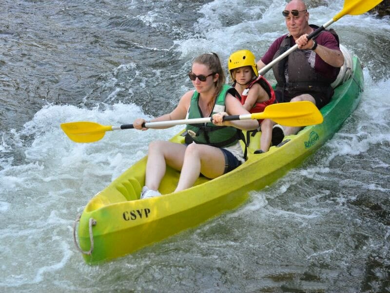 Trois personnes en kayak vert pagayant dans des rapides, avec un enfant portant un casque jaune.