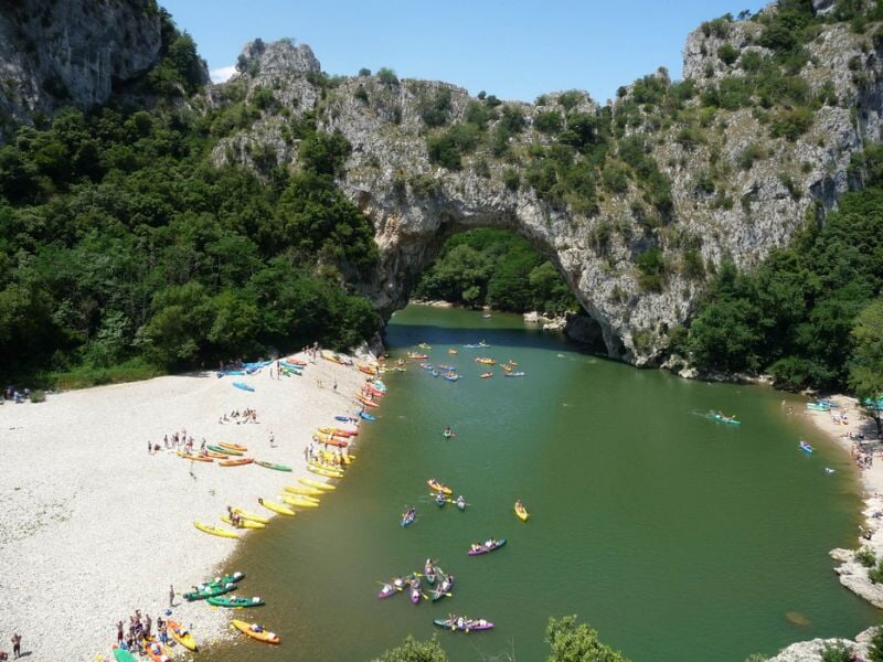 Vue aérienne d'une rivière avec des canoës colorés et des baigneurs près d'un pont naturel en pierre.