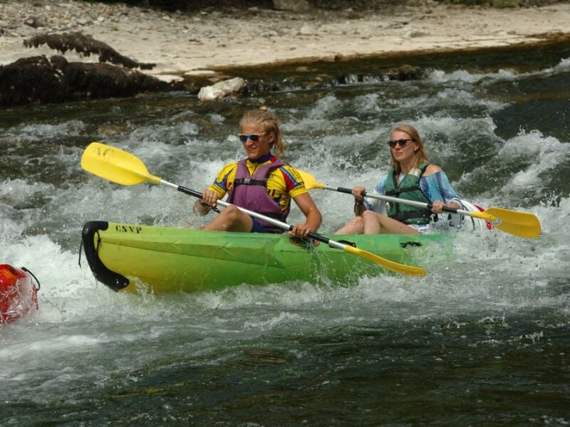Deux personnes pagayant en kayak vert sur une rivière avec des rapides et une ambiance ensoleillée.