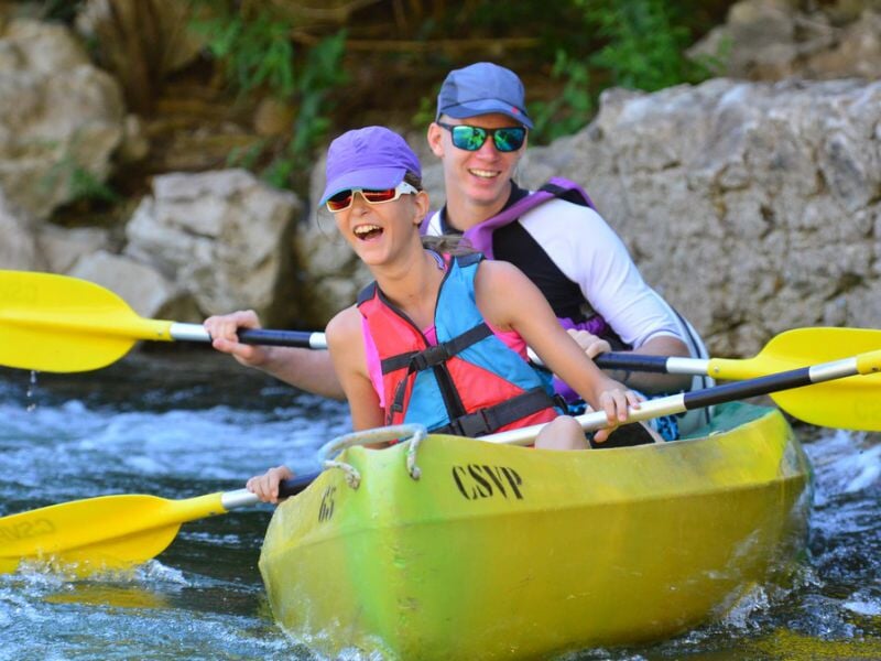 Un couple souriant pagayant ensemble dans un kayak jaune sur une rivière calme entourée de rochers.