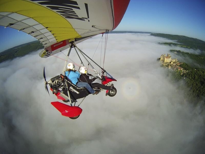 Un deltaplane motorisé avec deux passagers survolant un paysage enveloppé de brume avec un château médiéval émergeant des nuages