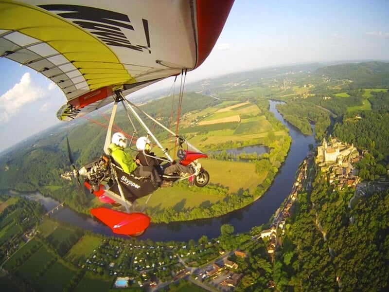 Un deltaplane motorisé avec deux passagers vole au-dessus d'un paysage pittoresque avec une rivière sinueuse et un château proche de la berge