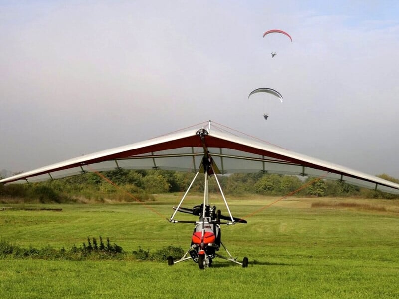 Un deltaplane motorisé décollant d'un champ avec deux parachutistes en arrière-plan dans le ciel
