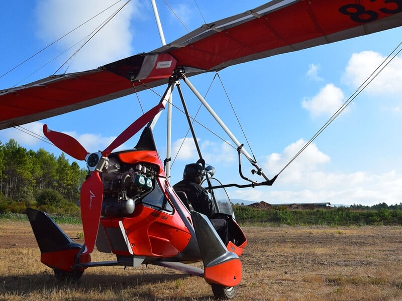 Un deltaplane motorisé rouge stationné sur un terrain herbeux, avec un pilote et un passager à bord, prêt pour un vol