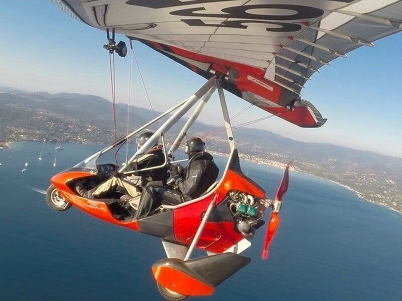 Vue en vol d'un deltaplane motorisé de couleur rouge avec deux personnes à bord, survolant une côte et une mer bleue avec des bateaux et une ville en arrière-plan