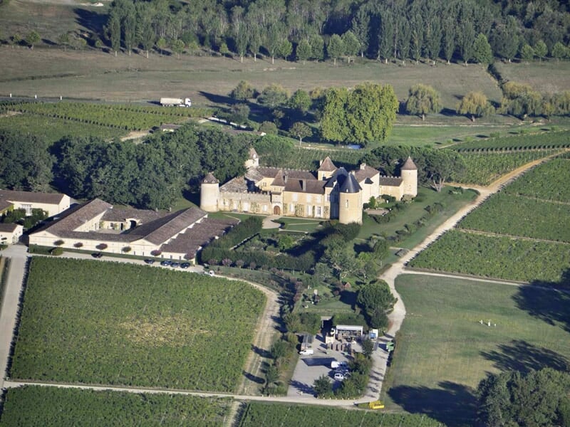 Vue aérienne d'une élégante demeure avec des tours, située dans un grand domaine agricole avec des rangées de vignes, indiquant une riche histoire dans la région de l'Entre deux mers