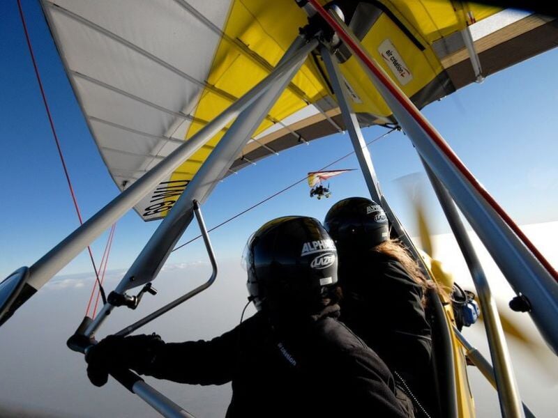 Deux personnes en tenue de vol, avec des casques, dans un deltaplane motorisé, avec une vue en contre-jour du ciel