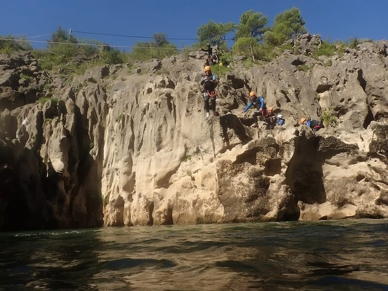 Personne en combinaison noire et casque rouge descendant une cascade rocheuse en canyoning.