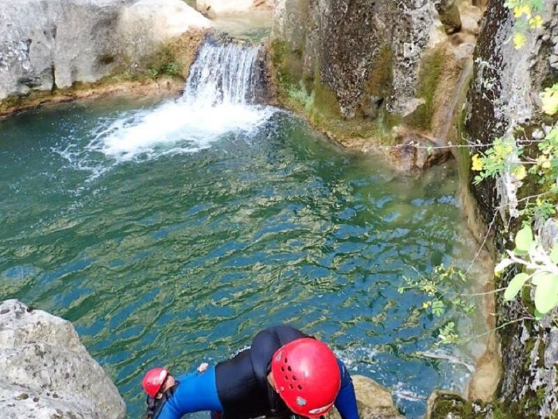 Personne en combinaison et casque rouge escaladant une roche au bord d'une cascade en canyoning.