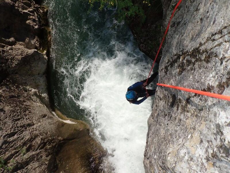 Canyoneur en combinaison bleue descendant en rappel le long d'une paroi rocheuse au-dessus d'une cascade.