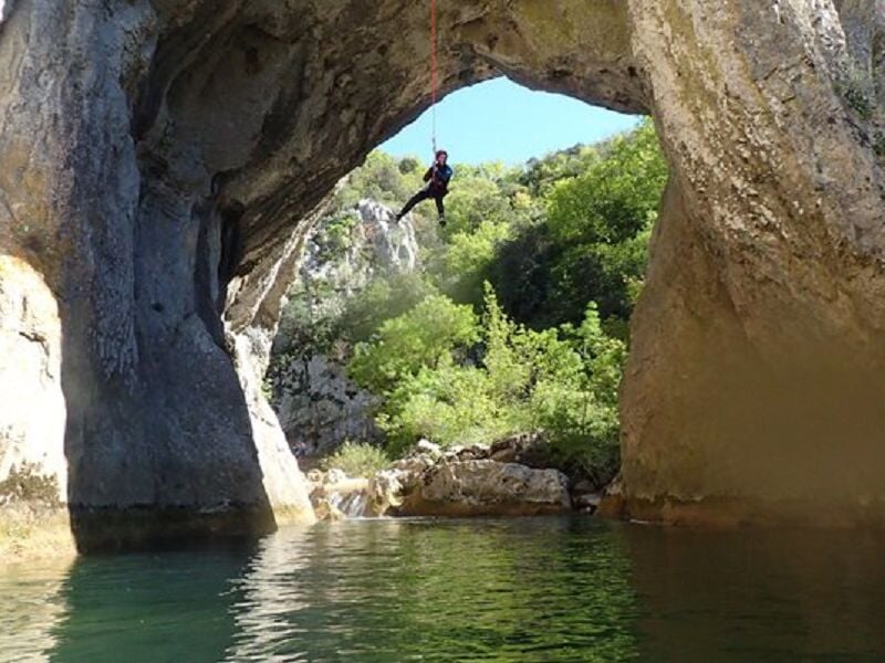Personne suspendue à une corde au-dessus d'une rivière sous un grand arc rocheux en pleine nature.