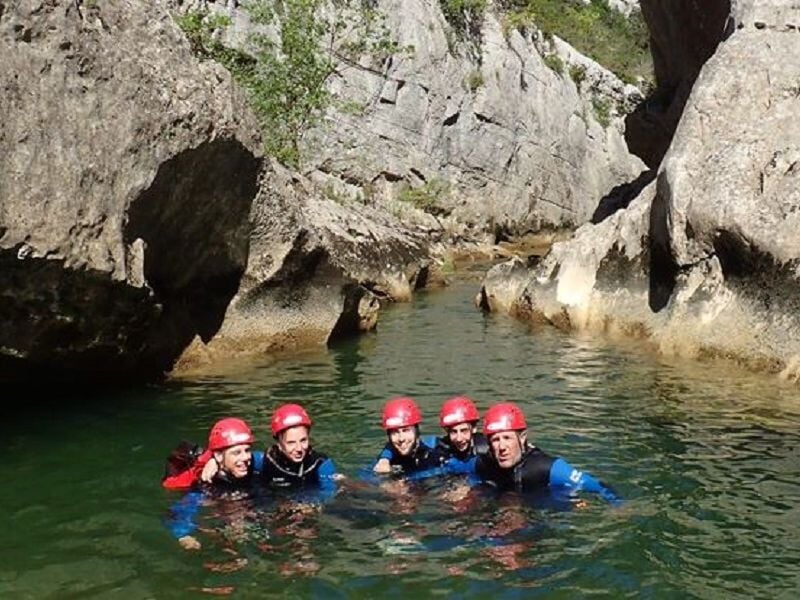 Groupe de personnes en combinaison et casque rouge nageant dans une rivière encaissée entre des falaises.