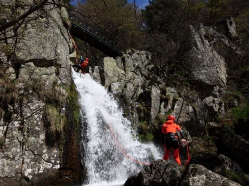Personne en combinaison rouge descendant en rappel une cascade dans un canyon rocheux et verdoyant.