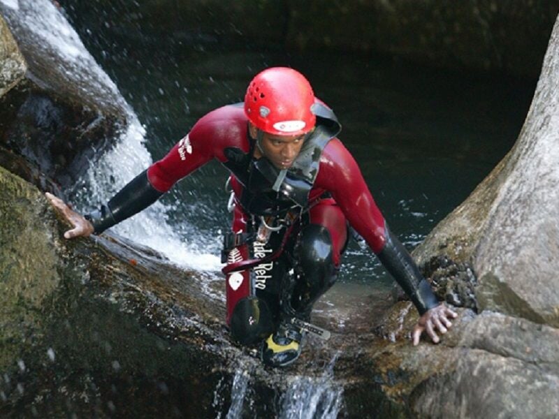 Personne en combinaison noire et casque rouge descendant une cascade rocheuse en canyoning.