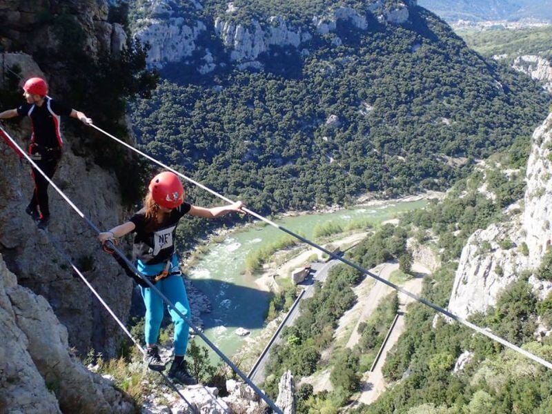 Personne en équipement de via ferrata traversant un pont suspendu avec vue sur une vallée.