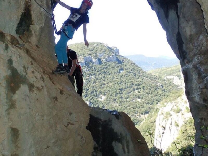 Personne en équipement de via ferrata progressant sur une paroi rocheuse avec vue panoramique.