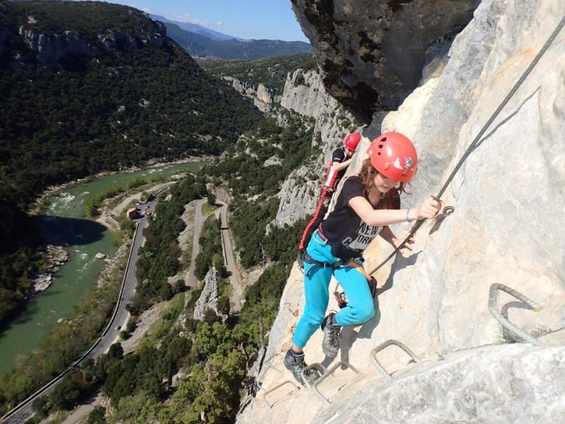 Personne en équipement de via ferrata escaladant une paroi rocheuse avec un paysage verdoyant.