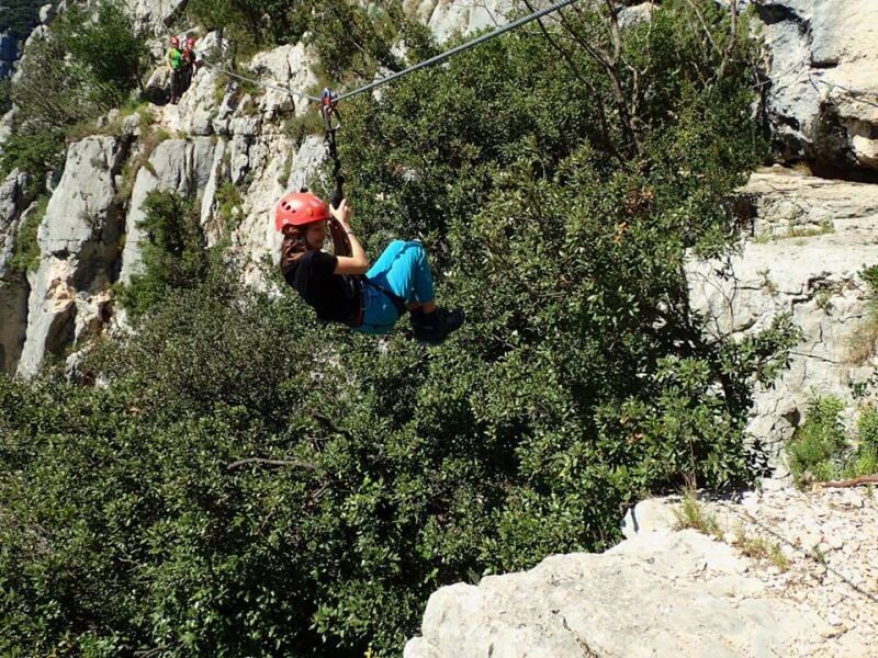 Personne en équipement de via ferrata escaladant une paroi rocheuse avec un paysage de vallée.