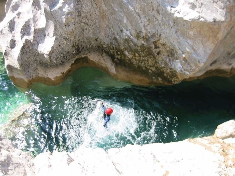 Personne en combinaison et casque rouge sautant dans une eau turquoise claire entourée de rochers.