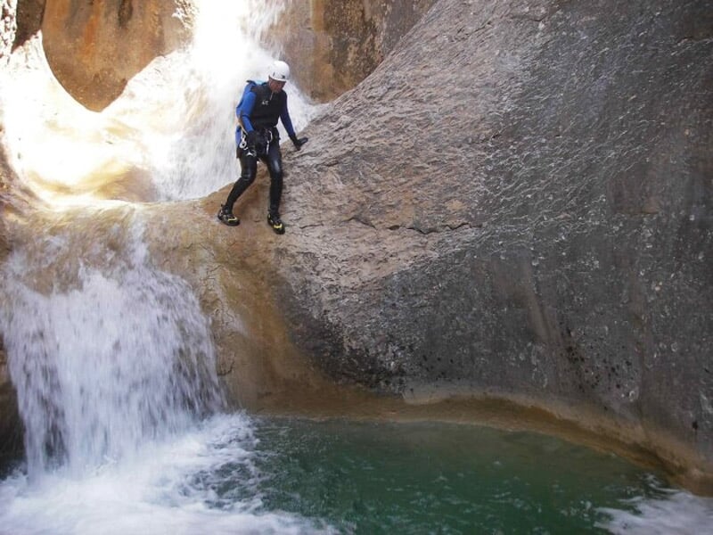 Personne en combinaison et casque bleu descend en rappel une cascade dans un canyon avec eau turquoise.
