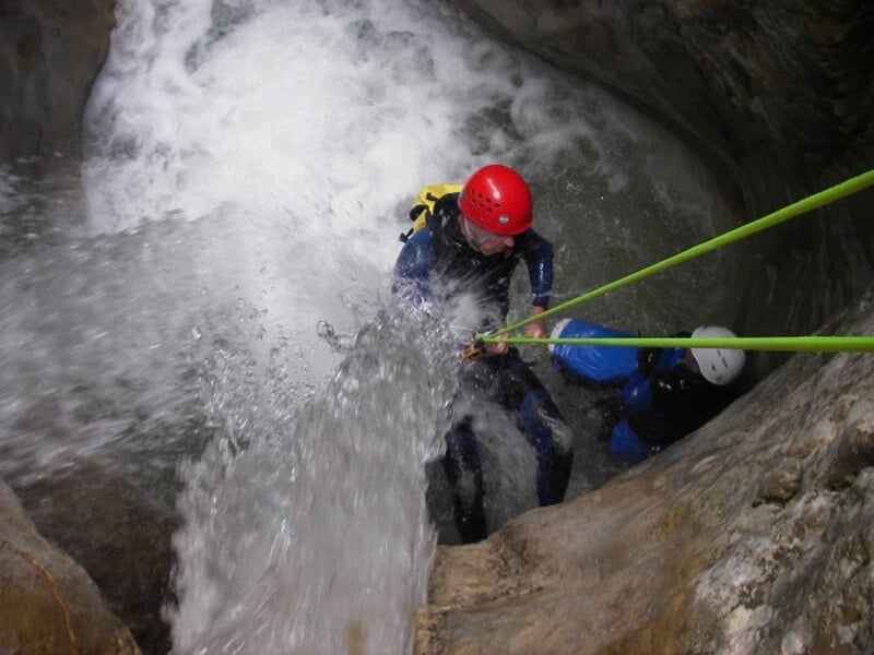Deux personnes en combinaison et casque descendent en rappel sous une cascade dans un canyon rocheux.