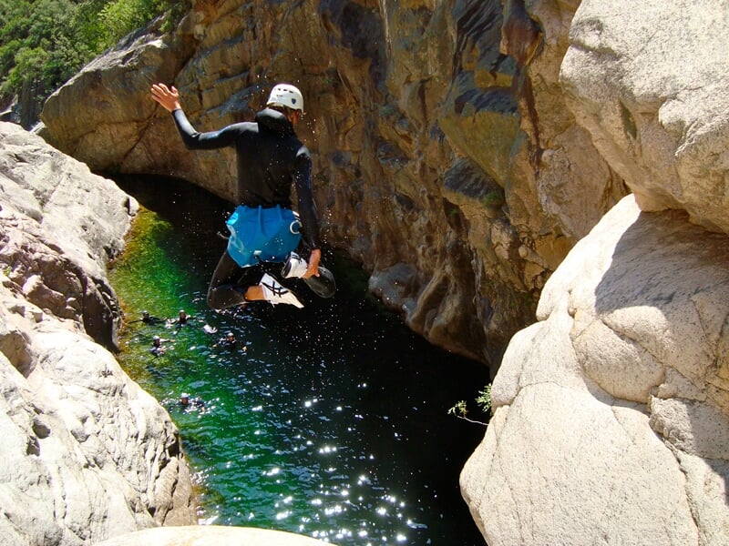 Personne en combinaison noire et casque blanc saute dans une eau claire entourée de rochers dans un canyon.