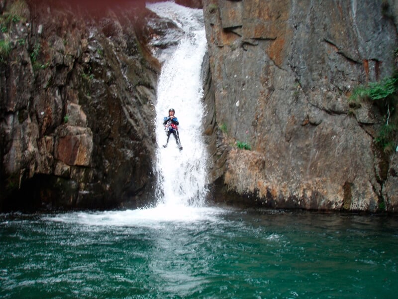 Personne en combinaison rouge descend en rappel une cascade dans un canyon rocheux avec eau en contrebas.