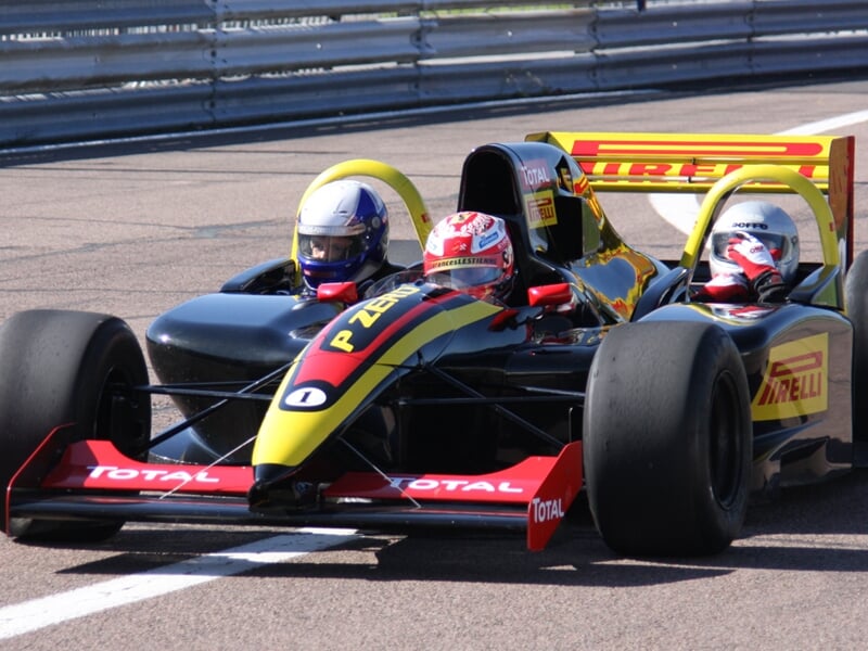 Deux pilotes casqués dans une monoplace noire et rouge en pleine course sur un circuit.