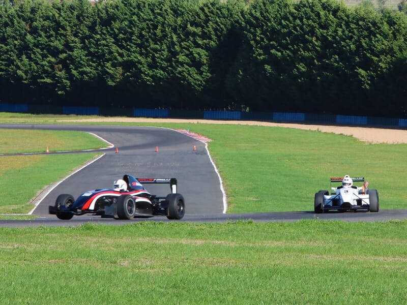 Deux monoplaces de Formule Renault en pleine course sur une piste entourée d'herbe verte et d'arbres au circuit.