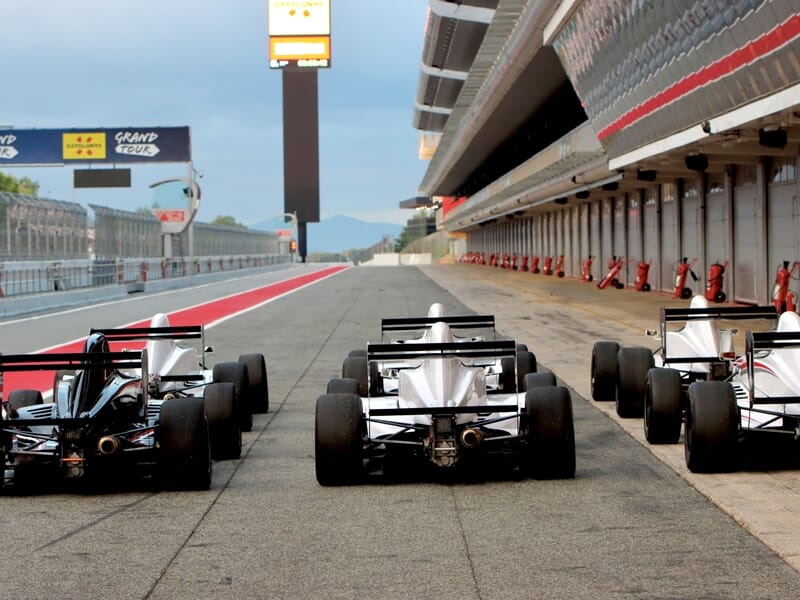 Quatre monoplaces alignées dans la voie des stands d'un circuit de course sous un ciel nuageux.