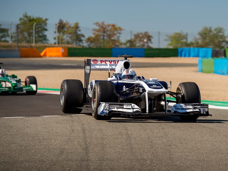 Formule 1 Williams bleue et blanche en virage sur le circuit de Magny-Cours avec un autre bolide vert en arrière-plan.