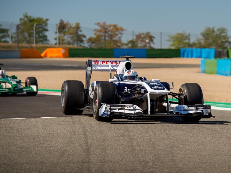 Formule 1 Williams bleue et blanche en virage sur le circuit de Magny-Cours avec un autre bolide vert en arrière-plan.